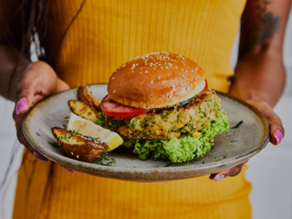 A woman holds a healthy vegan burger on a handmade ceramic plate, made of zucchini, green pea, seasoning, herbs and spices, close up